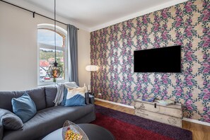 En stue med en sofa, et fladskærms-tv monteret på væggen, et vindue med gardiner og tapet med blomstermønster.