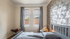 A bedroom with a bed, a radiator, a window with curtains, a chair, and a wall with a floral pattern. - Apartment 'Residenz Unter Der Birke - Esche' with Mountain View, Shared Terrace and Wi-Fi (Eisenach)