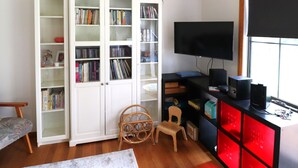Ein Zimmer mit einem weißen Bücherregal, einem Fernseher, einem Schreibtisch mit Computer und einem Fenster.