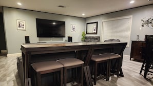 Cozinha moderna com ilha central, banquetas, televisão de tela plana e janela com persianas.