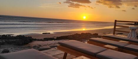 A beachside lounge with wooden benches and cushions, overlooking the ocean at sunset.