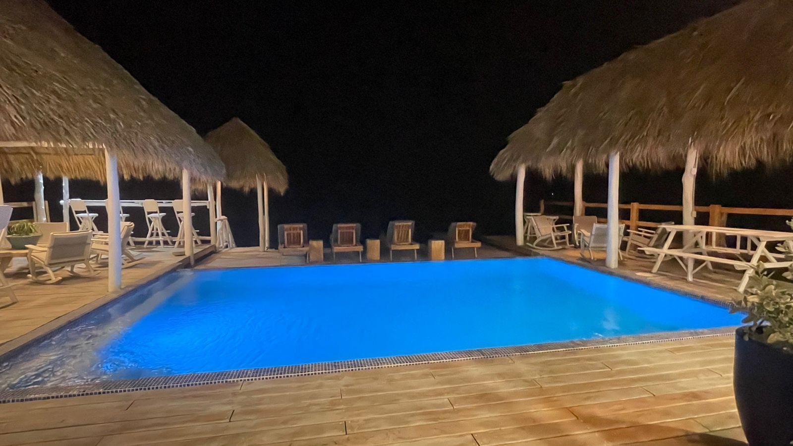 A swimming pool with thatched roof umbrellas, lounge chairs, and wooden decking.