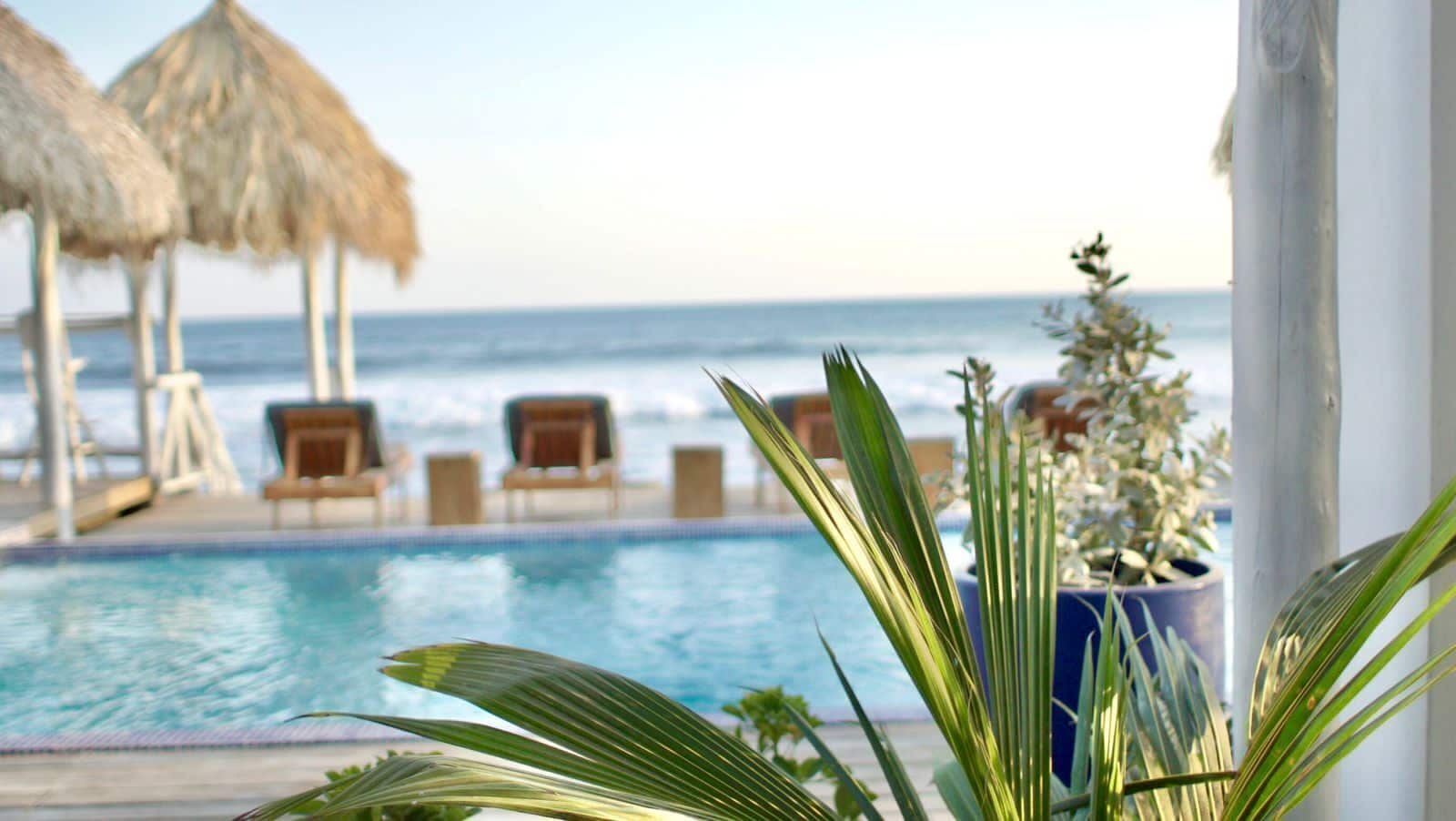 A poolside view with a clear view of the ocean, lounge chairs, and thatched umbrellas.