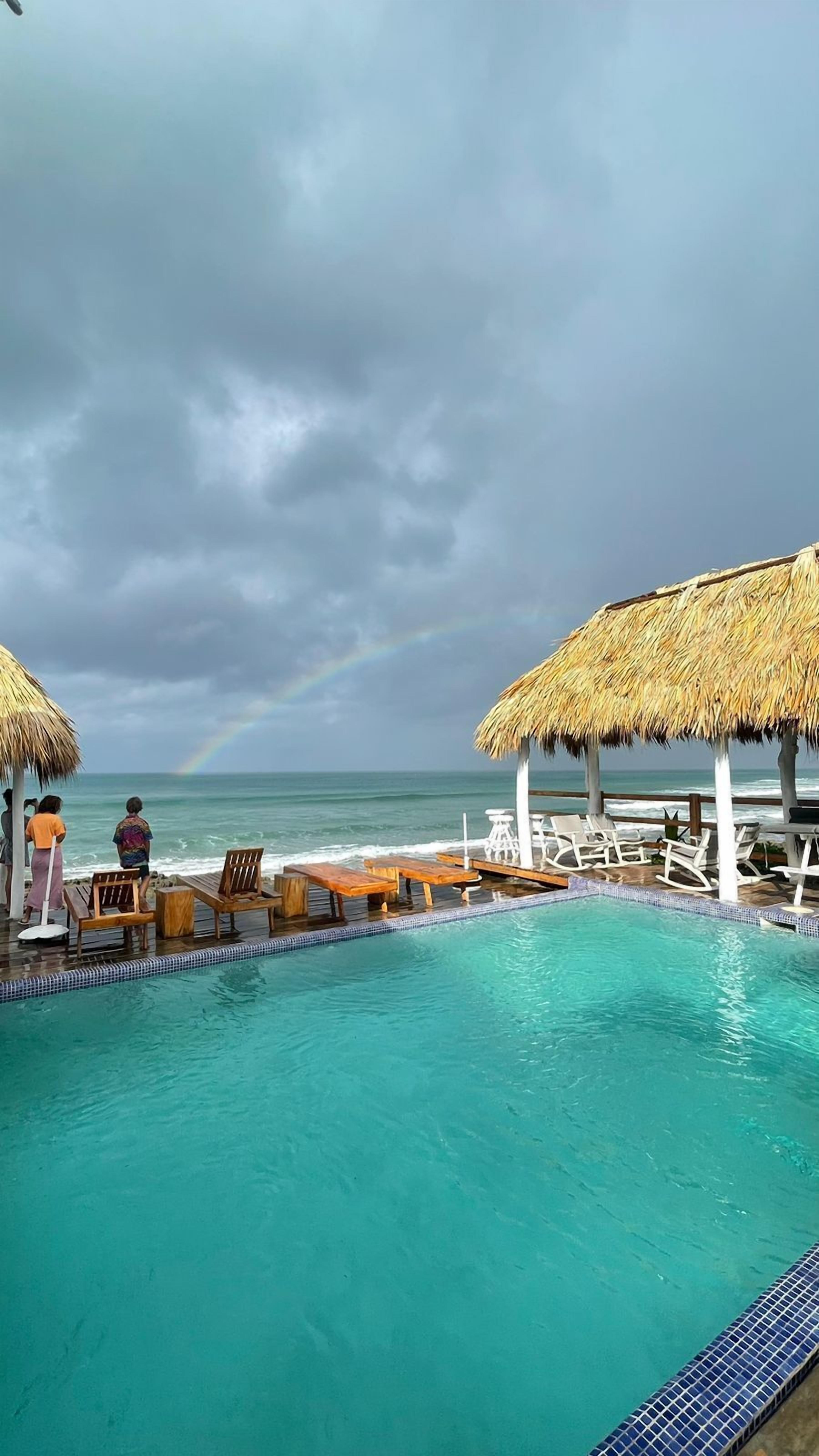 A poolside area with lounge chairs and a thatched umbrella, overlooking the ocean with a visible rainbow.