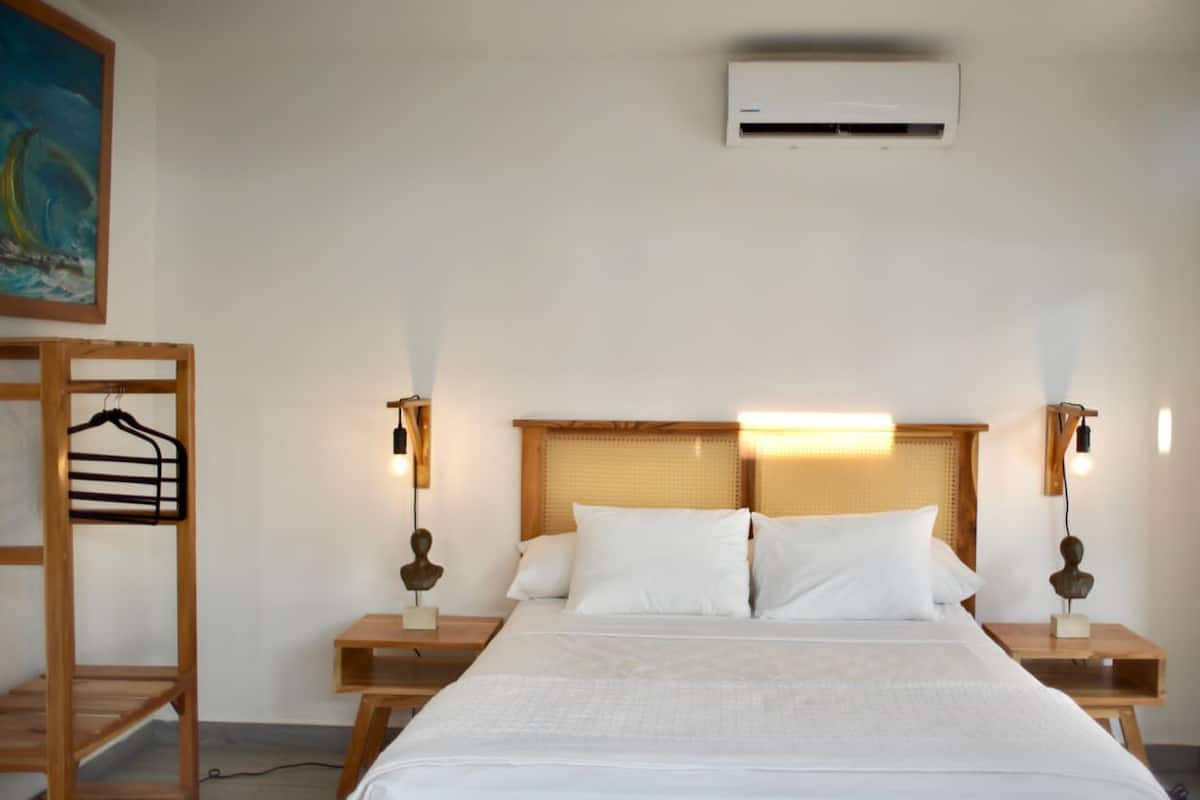 A hotel room with a large bed, wooden headboard, two bedside tables, a wall-mounted air conditioner, and a painting on the wall.