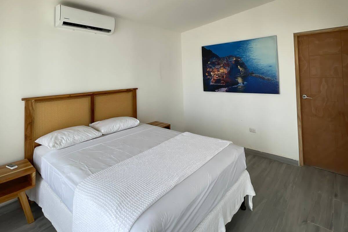 A bedroom with a large bed, a wooden headboard, a nightstand, an air conditioning unit, a painting, and a door.