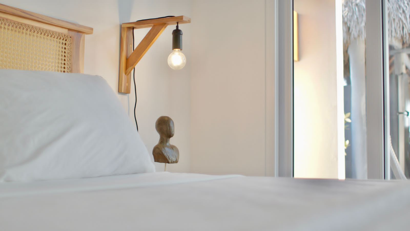 A bed with white linens, a wooden headboard, a wall-mounted light fixture, and a bust sculpture.