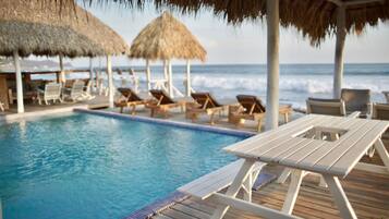 Un área junto a la piscina con sillones de descanso y sombrillas de paja, con vista al océano.