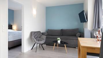 Une pièce moderne avec un canapé gris, une petite table ronde, un fauteuil gris, un téléviseur fixé au mur et un lit dans la pièce adjacente.
