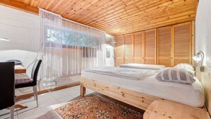 A bedroom with a wooden ceiling, a bed with a white mattress, a chair, a desk, and a window with sheer curtains.