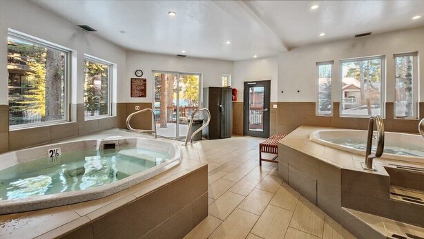Un área de spa con dos jacuzzis, un banco y amplios ventanales con vista a árboles.