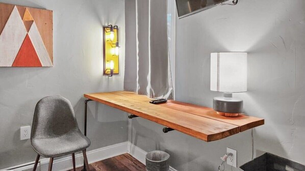 Une chambre moderne avec un bureau en bois, une chaise grise, une lampe fixée au mur et un tableau aux motifs géométriques.