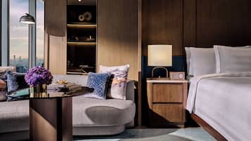 Habitación de hotel con cama, mesita de noche, mesa con lámpara, sofá con cojines, un jarrón con flores y vistas a la ciudad a través de amplias ventanas.