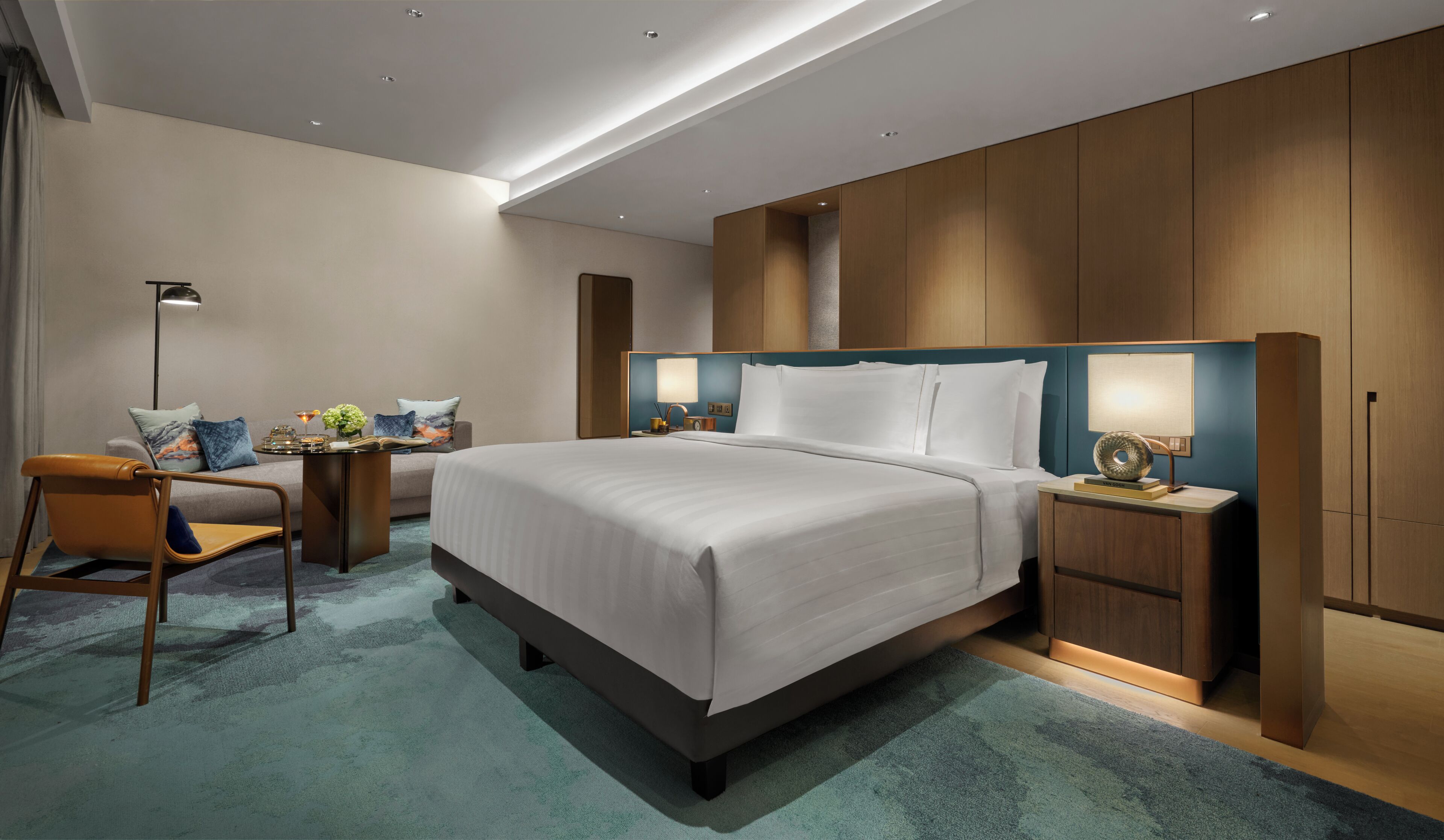 A modern hotel room with a large bed, a desk, a chair, and a nightstand with a lamp.