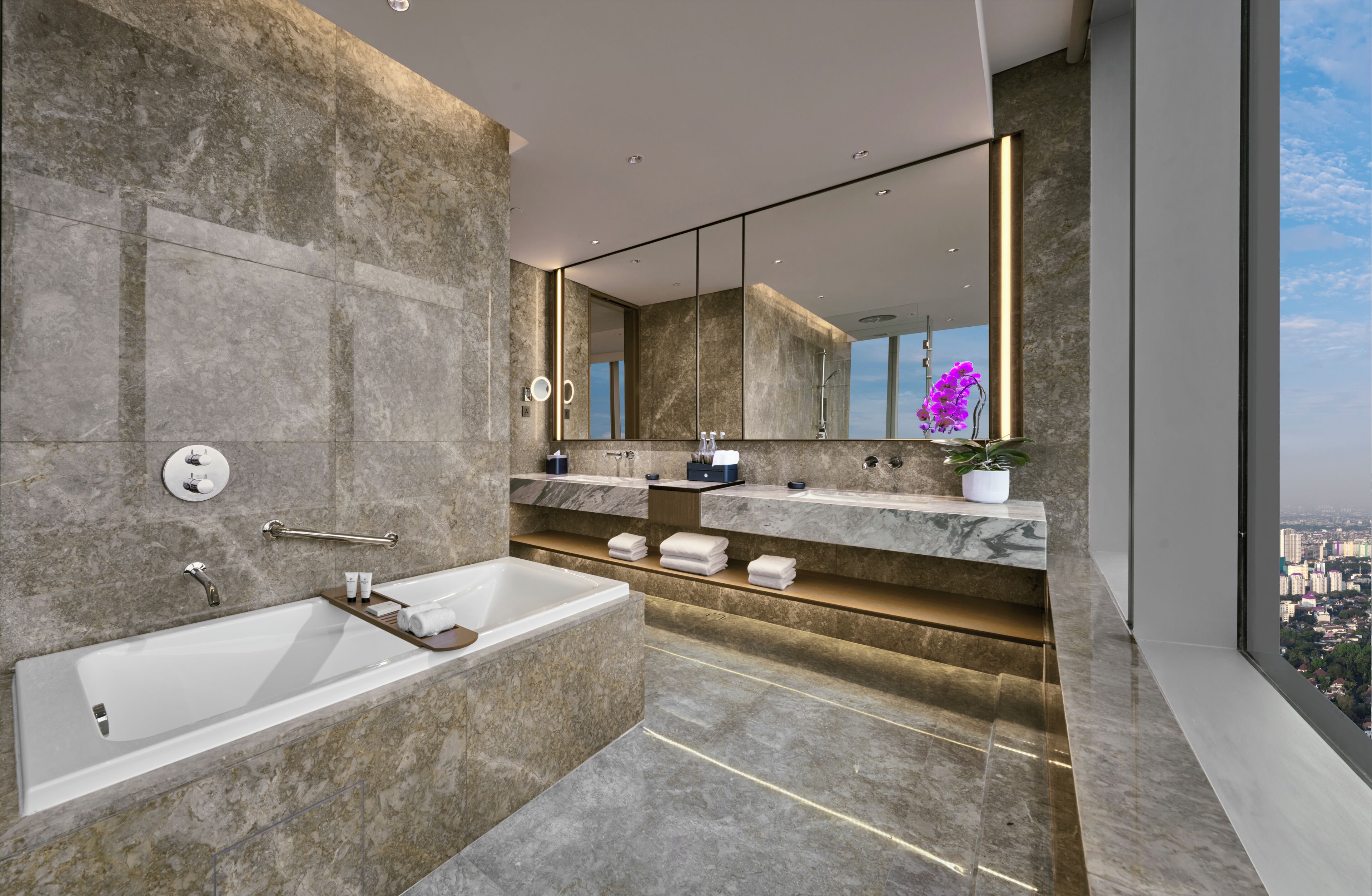 A modern bathroom with a large bathtub, a vanity with a mirror, and a view of the city.