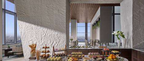 Un desayuno bufé de alta gama con una variedad de pasteles, frutas y bebidas, en un interior moderno con ventanas amplias y vistas a la ciudad.