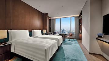 A hotel room with two beds, a desk, a chair, a TV, and a large window with a city view.