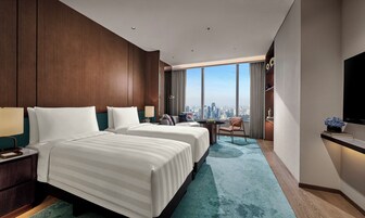 A hotel room with two beds, a desk, a chair, a TV, and a large window with a city view.
