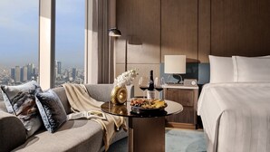 Habitación de hotel con vista a la ciudad, una cama, un sofá, una mesita con una botella de vino y copas, y una lámpara.