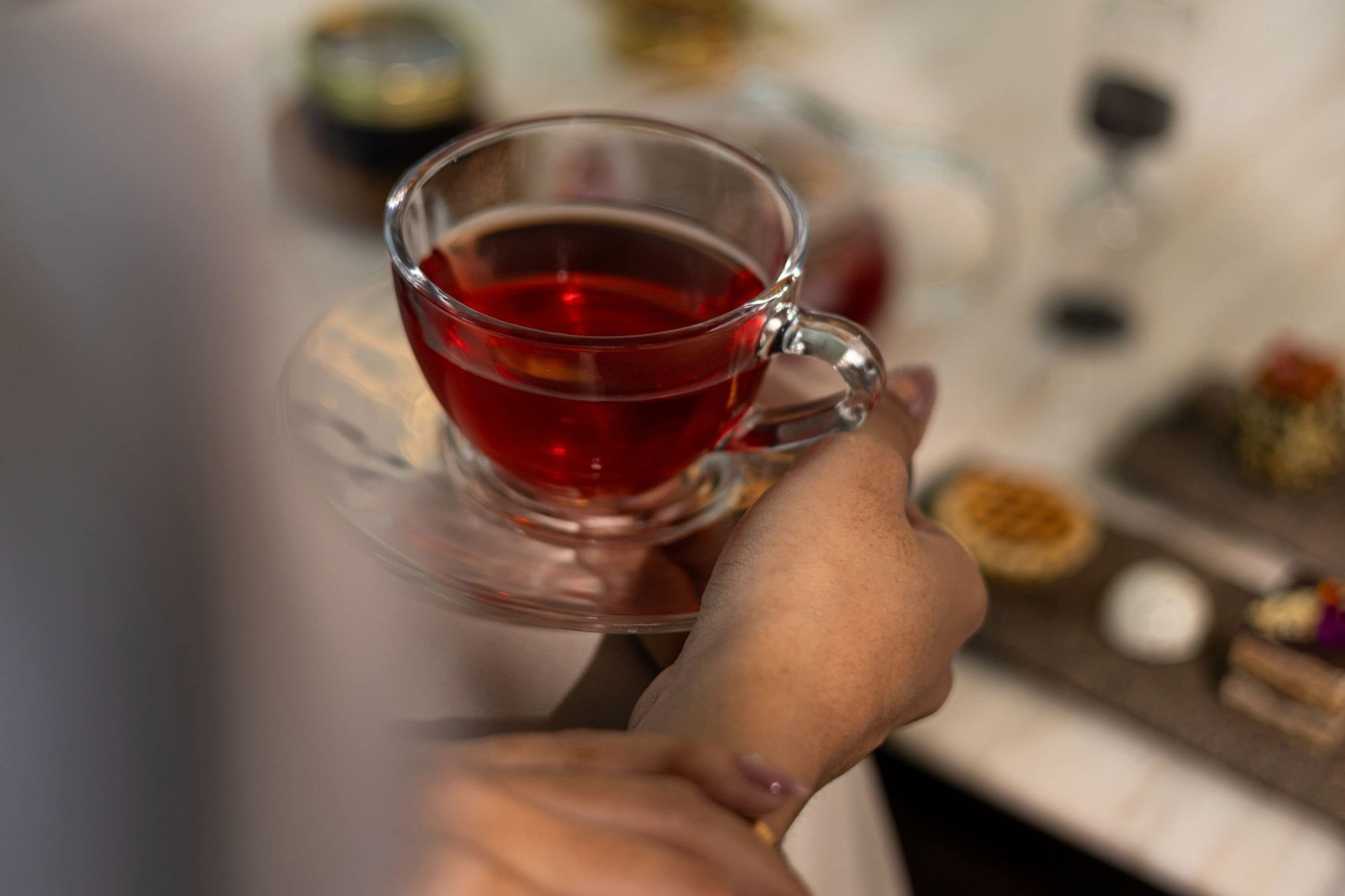 A hand holding a clear glass cup with a red liquid, possibly tea, on a saucer.