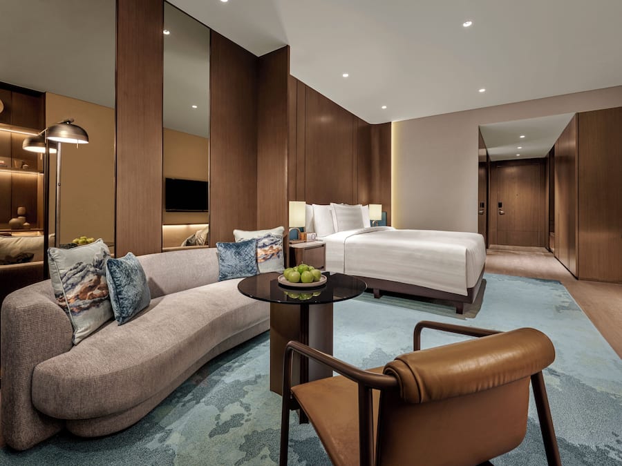 Quarto de hotel moderno com sofá curvo, mesinha de centro redonda, uma cama de solteiro, pequena mesa com frutas e televisão fixada na parede.