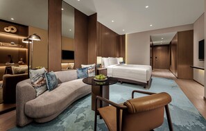 A modern hotel room with a curved sofa, a round coffee table, a single bed, a small table with fruit, and a TV mounted on the wall. - Pan Pacific Jakarta (Jakarta)