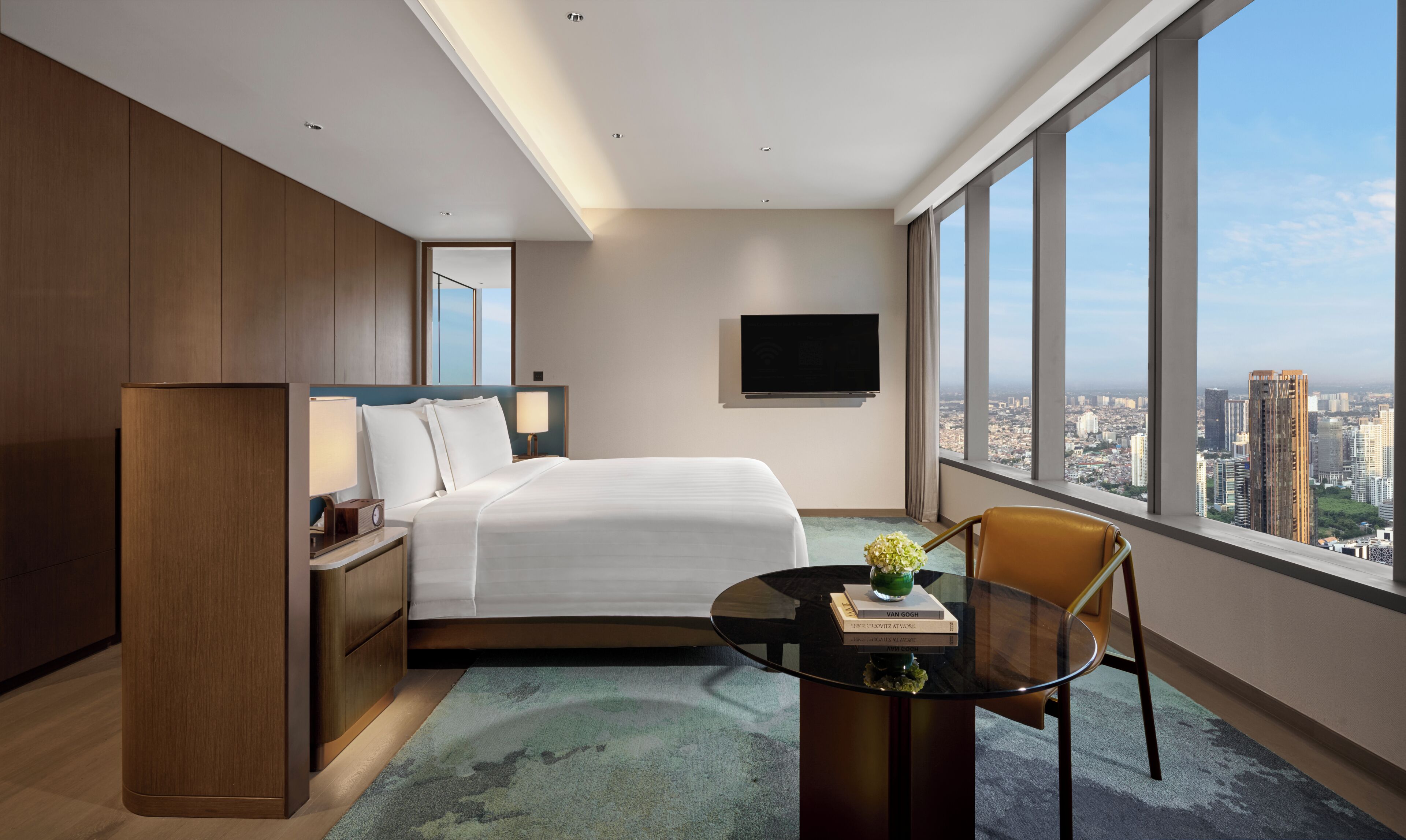 Una habitación de hotel moderna con una cama grande, un escritorio con una silla, un televisor montado en la pared y vistas a la ciudad a través de amplios ventanales.