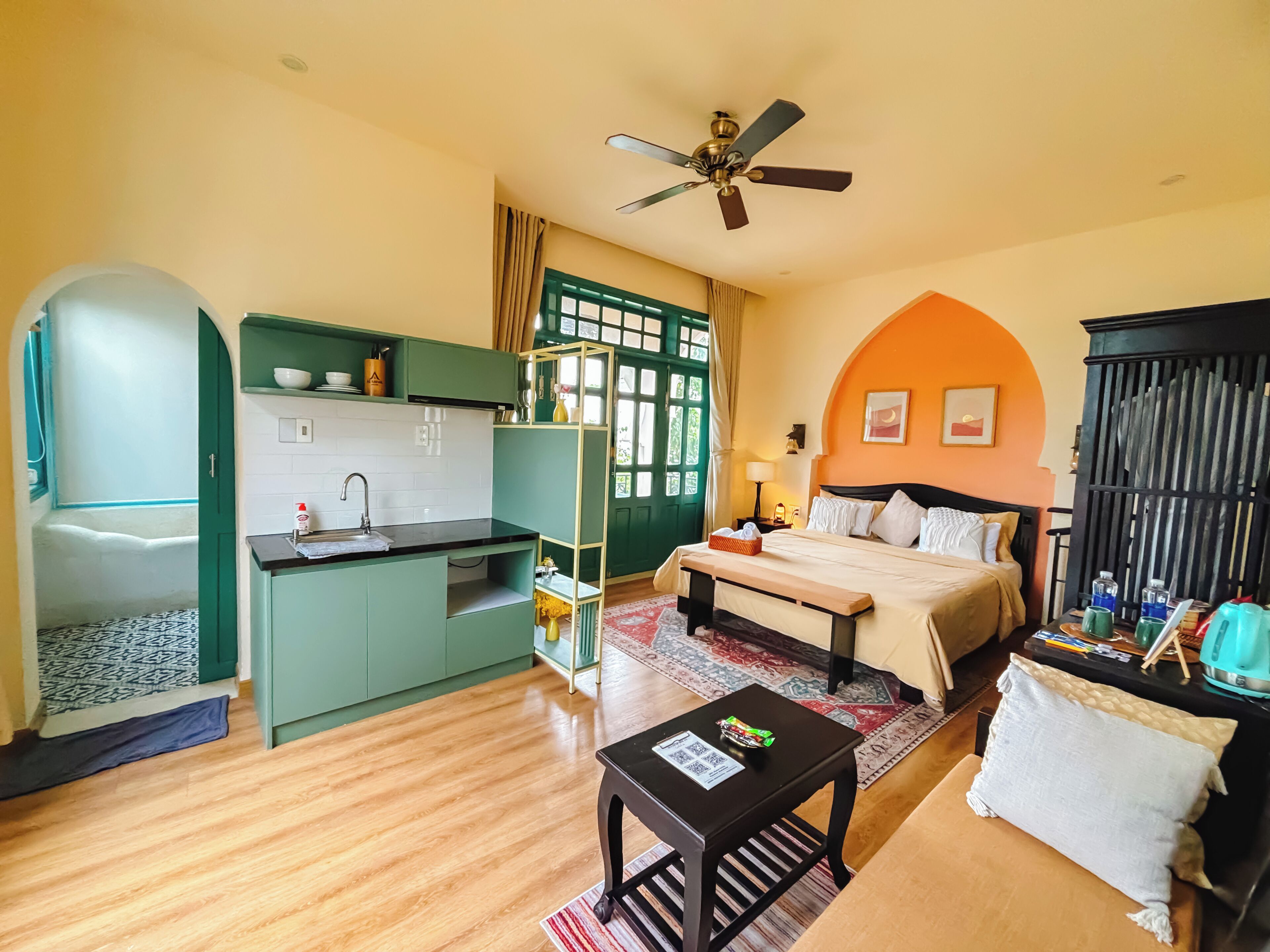 Une chambre d’hôtel compacte comprenant un coin cuisine, un lit avec une tête de lit, une petite table et un ventilateur de plafond.