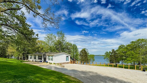 A single-story house with a gravel driveway leading to a wooden deck, surrounded by lush greenery and a lake view. - KY Lakeshore Retreat -Waterfront Views, Dock, Deck, Large Parking -NEW 2024 (New Concord)