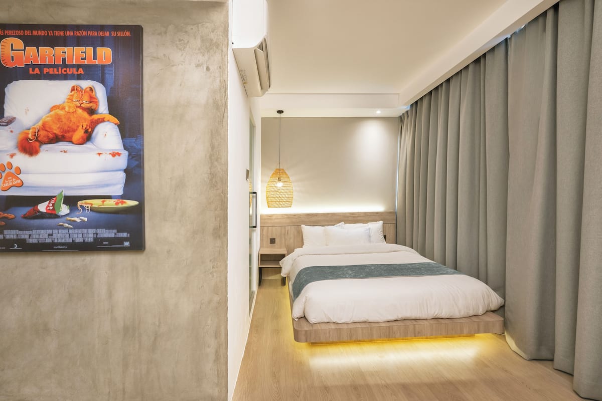 A hotel room with a bed, a poster of Garfield, a wall-mounted air conditioner, and a bedside lamp.