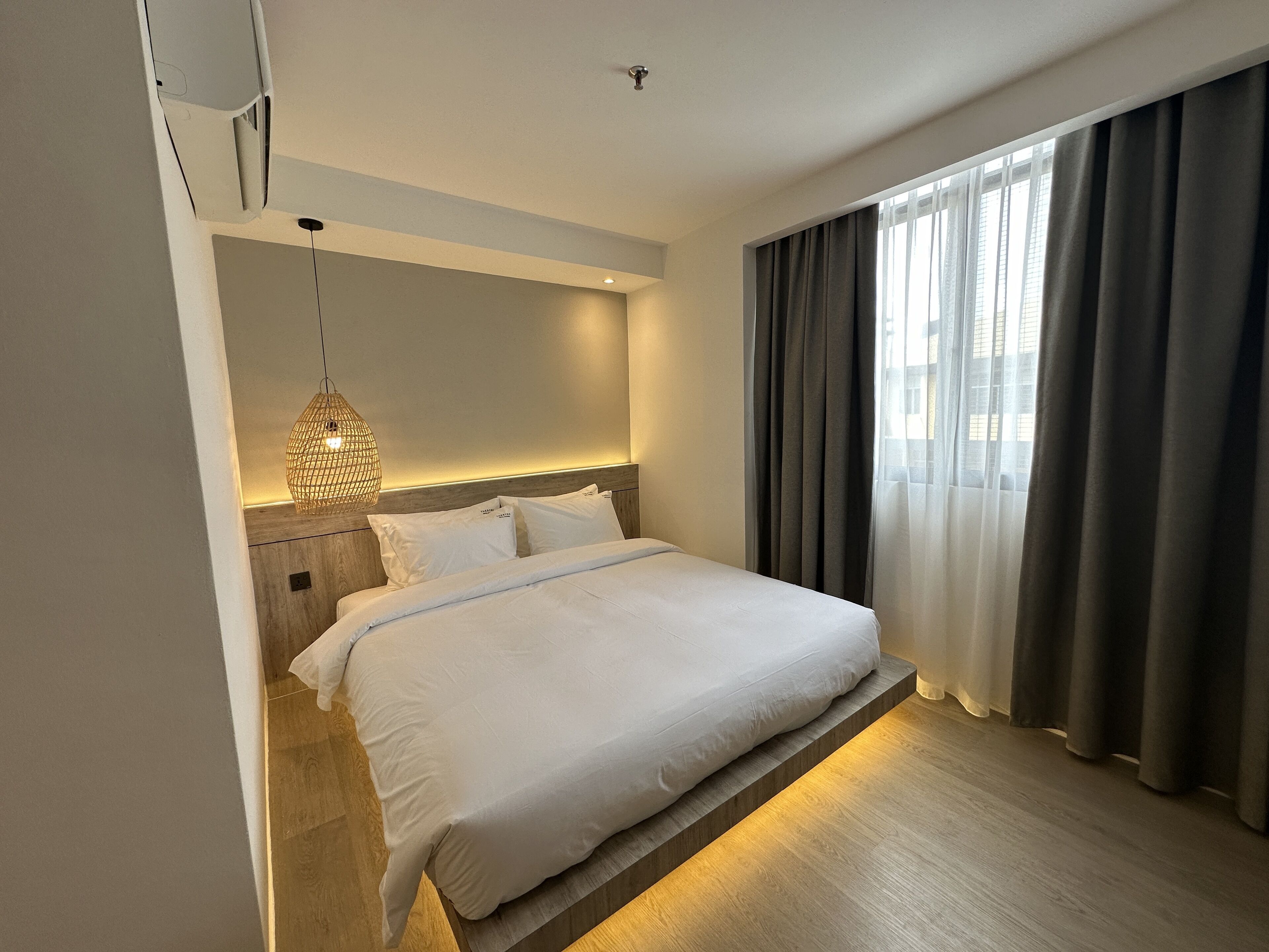 A modern hotel room with a neatly made bed, a hanging light fixture, and a view of the city through large windows.