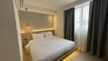 A modern hotel room with a neatly made bed, a hanging light fixture, and a view of the city through large windows.