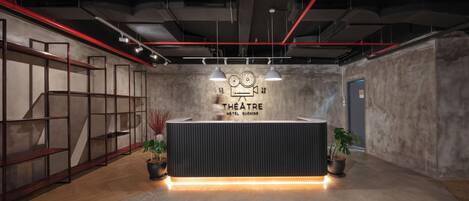 En receptionsdisk med et skilt der siger "THEATRE HOTEL KUCHING" og et moderne interiør med en støbt gulv og metalkasser.