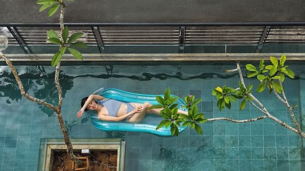 Una persona recostada en un flotador inflable en una piscina, con un árbol y una pequeña jardinera.