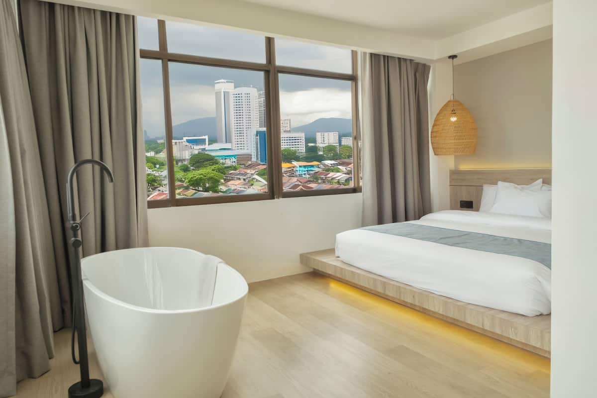 A hotel room with a large window, a bed, a white bathtub, and a hanging light fixture.