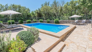 Une piscine rectangulaire bordée de pierres, entourée de chaises longues et de parasols, avec en arrière-plan des arbres et un mur de pierre.