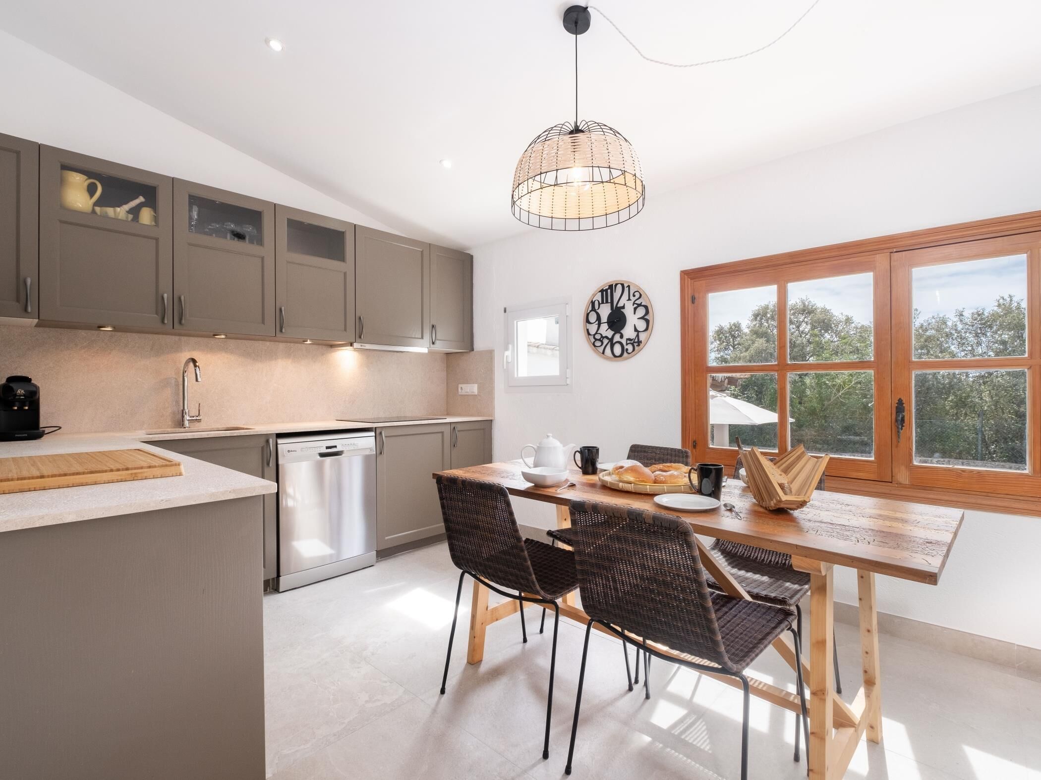 A modern kitchen with grey cabinets, a stainless steel dishwasher, and a wooden dining table with wicker chairs.