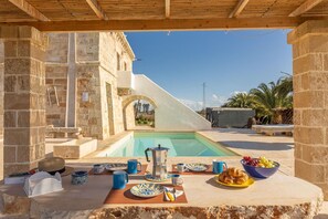 Colazione a bordo piscina con una moka, piatti e una ciotola di frutta.