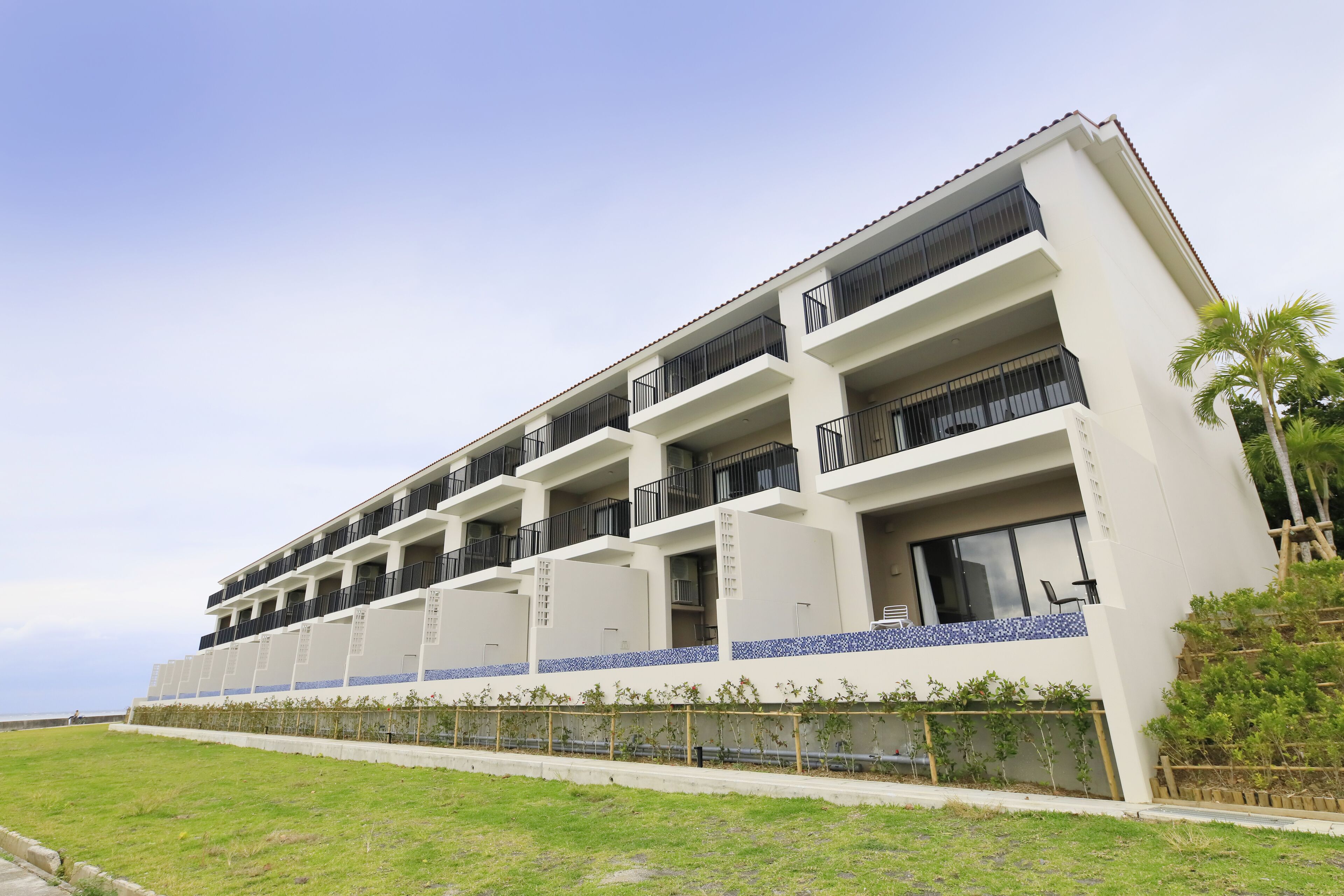 Un hotel de varios pisos con balcones, un nivel inferior con tejas y un área verde al frente.