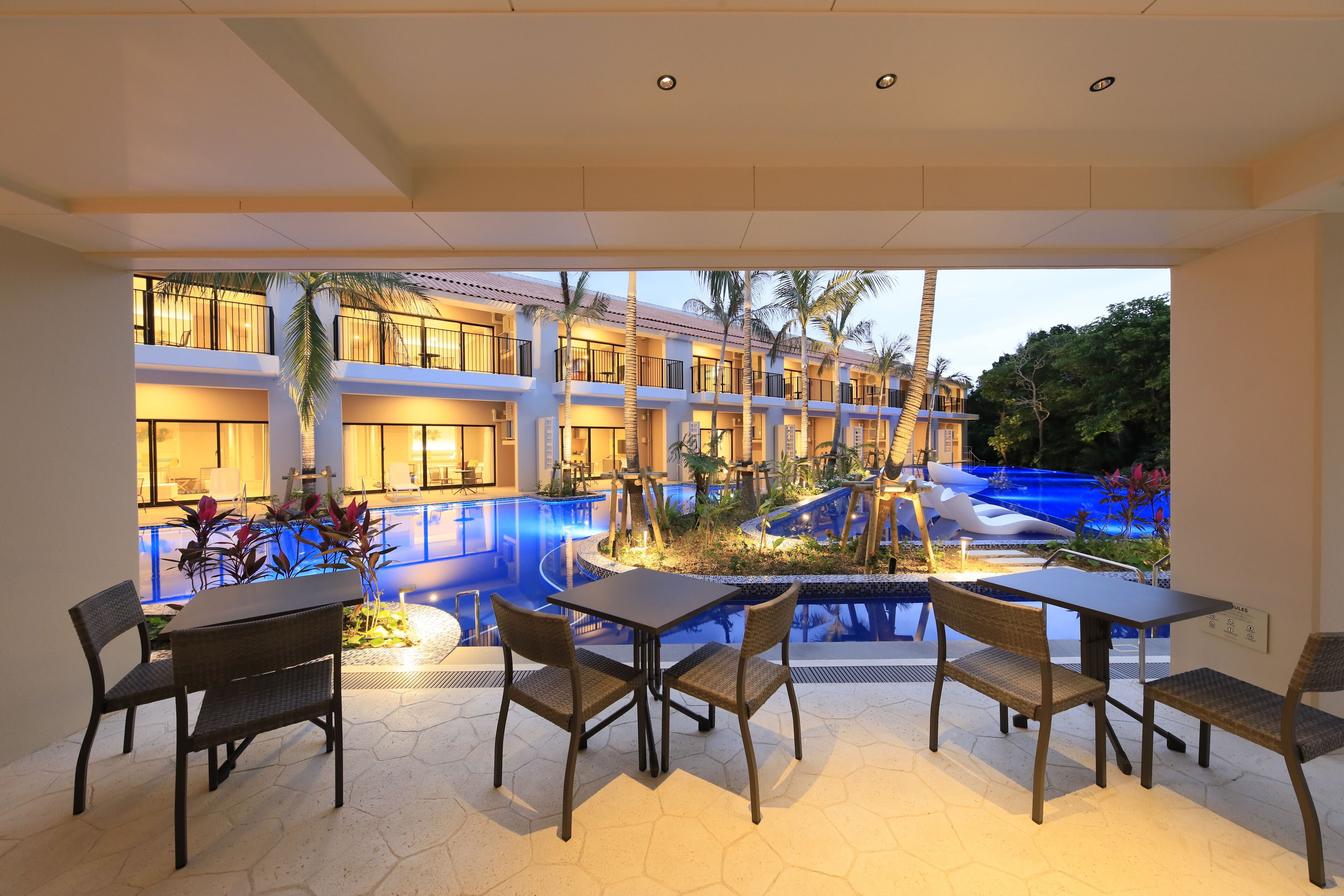 Vestíbulo de hotel con piscina, mobiliario exterior y vistas al área de la piscina.