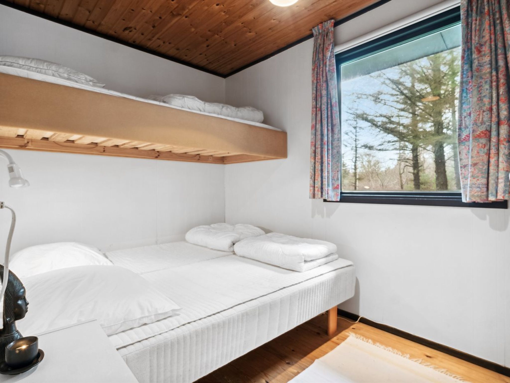 Un lit simple avec du linge de lit blanc, un plafond en bois, une fenêtre ornée de rideaux à motifs floraux et une petite table de chevet avec une lampe.