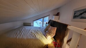 Un lit simple avec une tête de lit en bois, une fenêtre donnant sur des maisons et un tableau encadré accroché au mur.