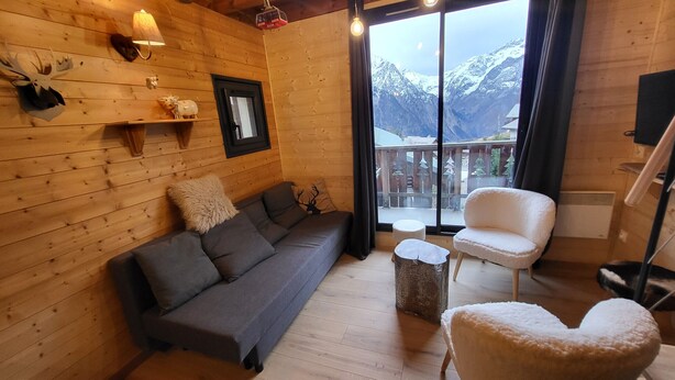 L’intérieur d’un chalet accueillant, avec un canapé, un fauteuil, une petite table et une fenêtre donnant sur les montagnes.