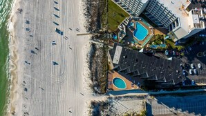 An aerial view of a coastal hotel with a beach, a swimming pool, and a parking area.