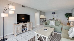 A modern living room with a flat-screen TV, a wooden entertainment unit, a light-colored coffee table, a green sofa, and a floor lamp.