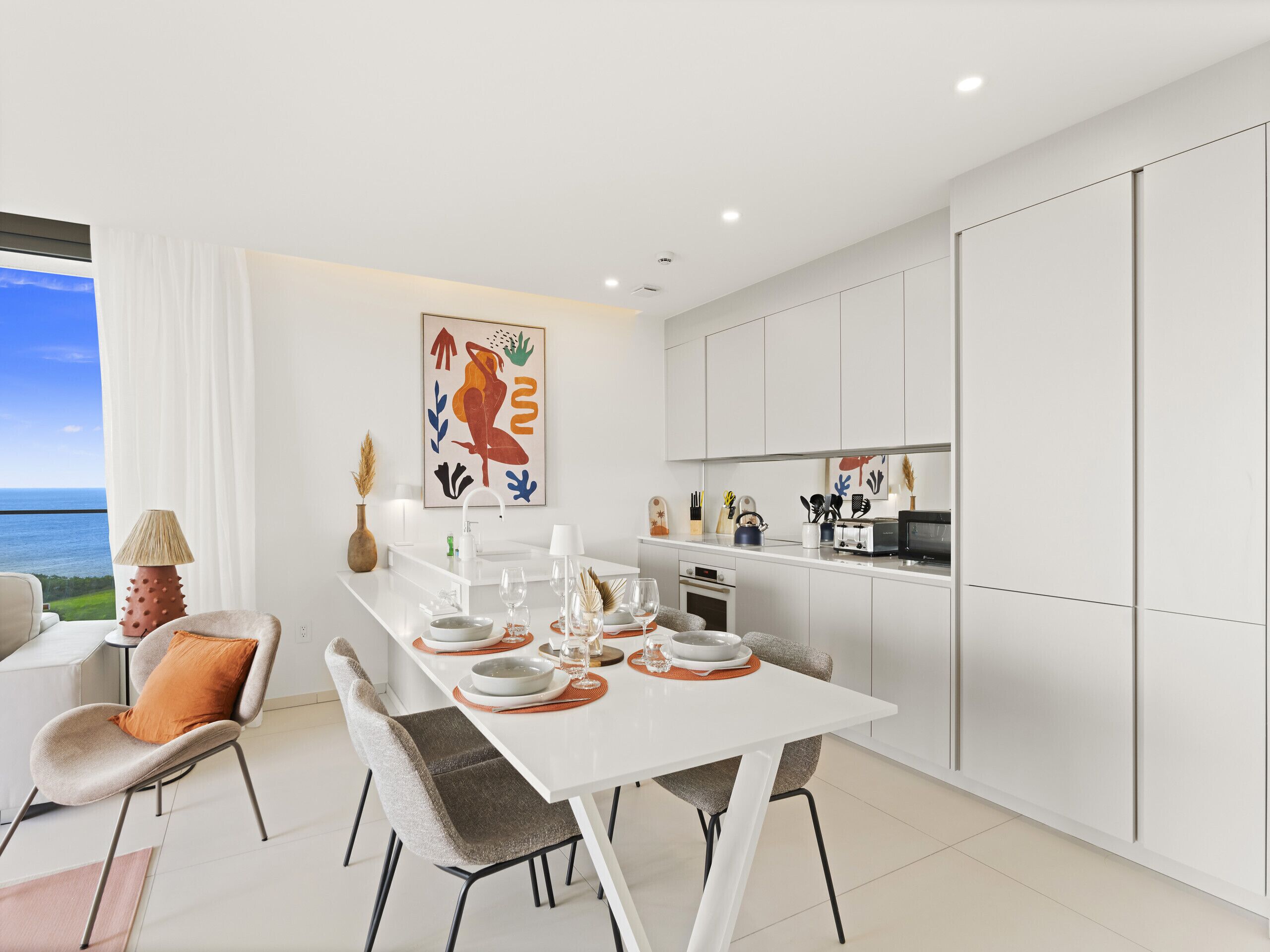 Een moderne keuken met witte kastjes, een eettafel voor vier personen en uitzicht op de oceaan door een groot raam.