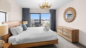 A hotel bedroom with a large bed, a wooden dresser, a round mirror, and a view of the city through a balcony.
