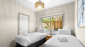 A hotel room with two single beds, a window with a view of greenery, a door, and a hanging light fixture.