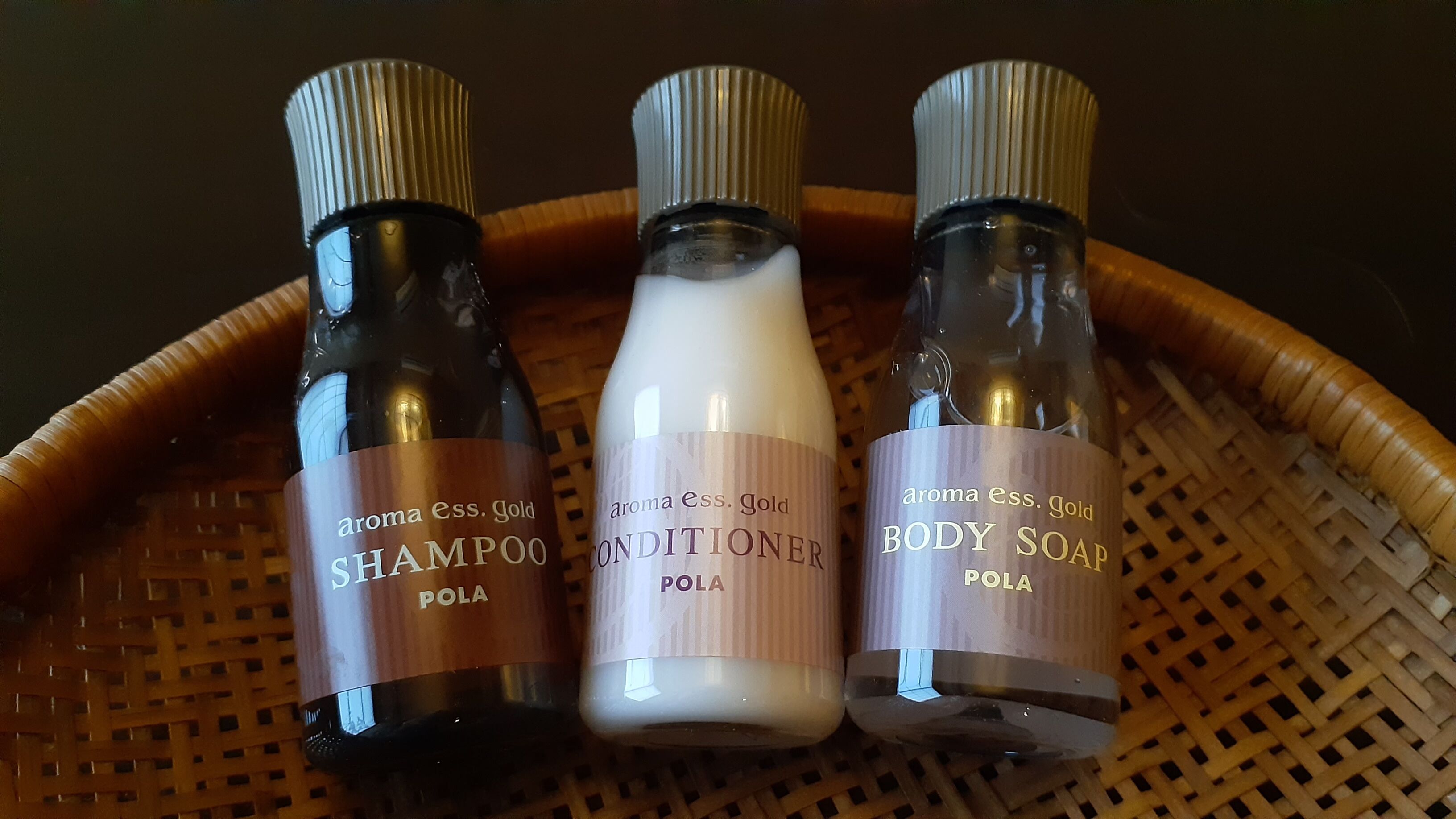 Três frascos de produtos 'Aroma Ess. Gold': 'Shampoo', 'Condicionador' e 'Sabonete Líquido', todos rotulados 'Pola', colocados numa cesta de palha.