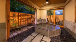 Un onsen japonés tradicional con interior de madera, un manantial termal de piedra y vista al bosque circundante a través de un puertas correderas.